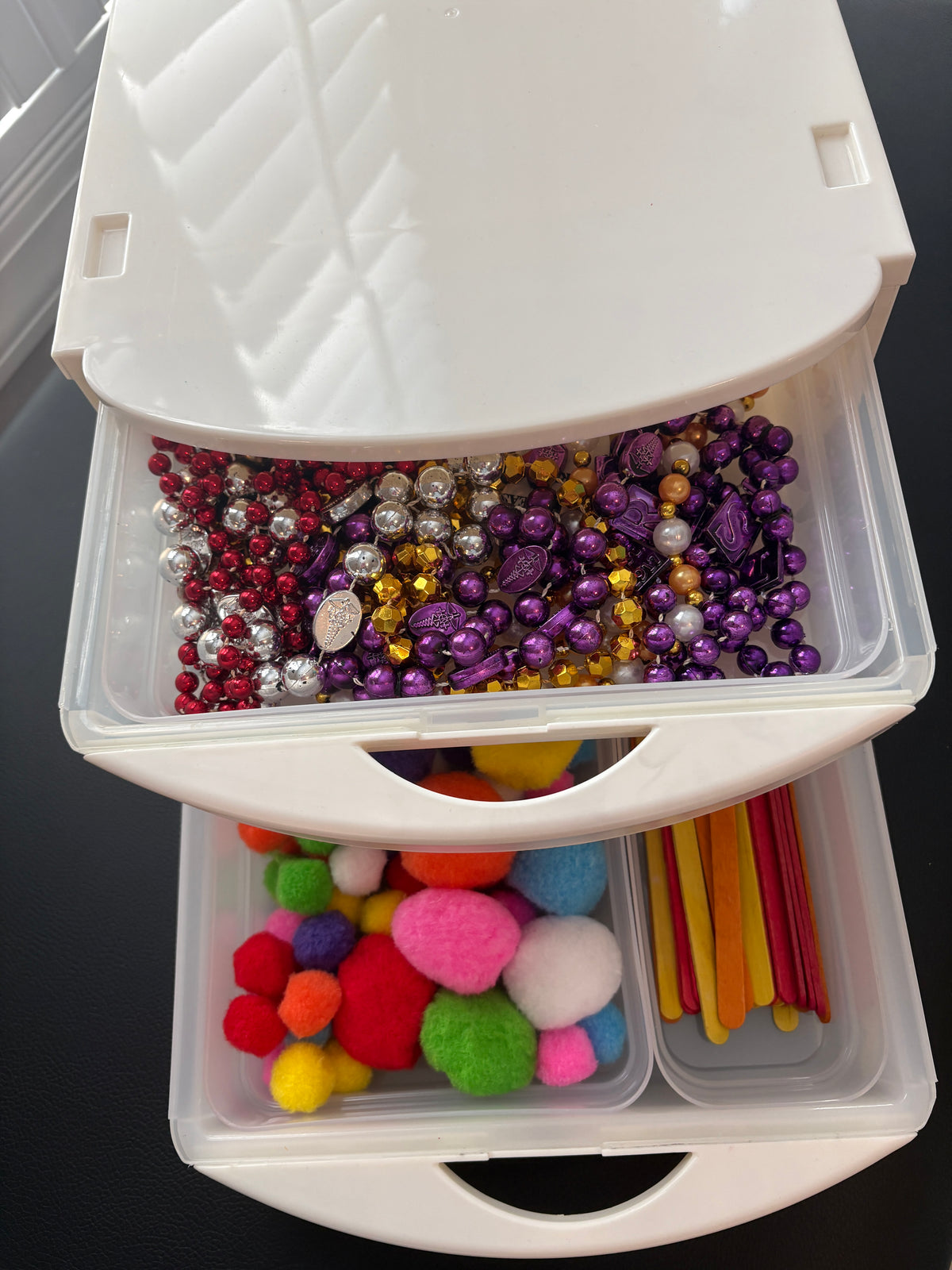 A white plastic drawer organizer with two open drawers filled with craft supplies, including colorful beads, pom-poms, and wooden sticks in slim modular removable 
compartments 