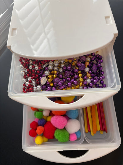 A white plastic drawer organizer with two open drawers filled with craft supplies, including colorful beads, pom-poms, and wooden sticks in slim modular removable 
compartments 