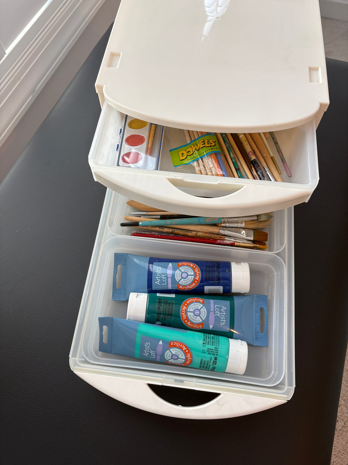 A white plastic multi-drawer storage organizer filled with art supplies, including paint tubes, brushes, and a small watercolor palette in slim-lined removable bins